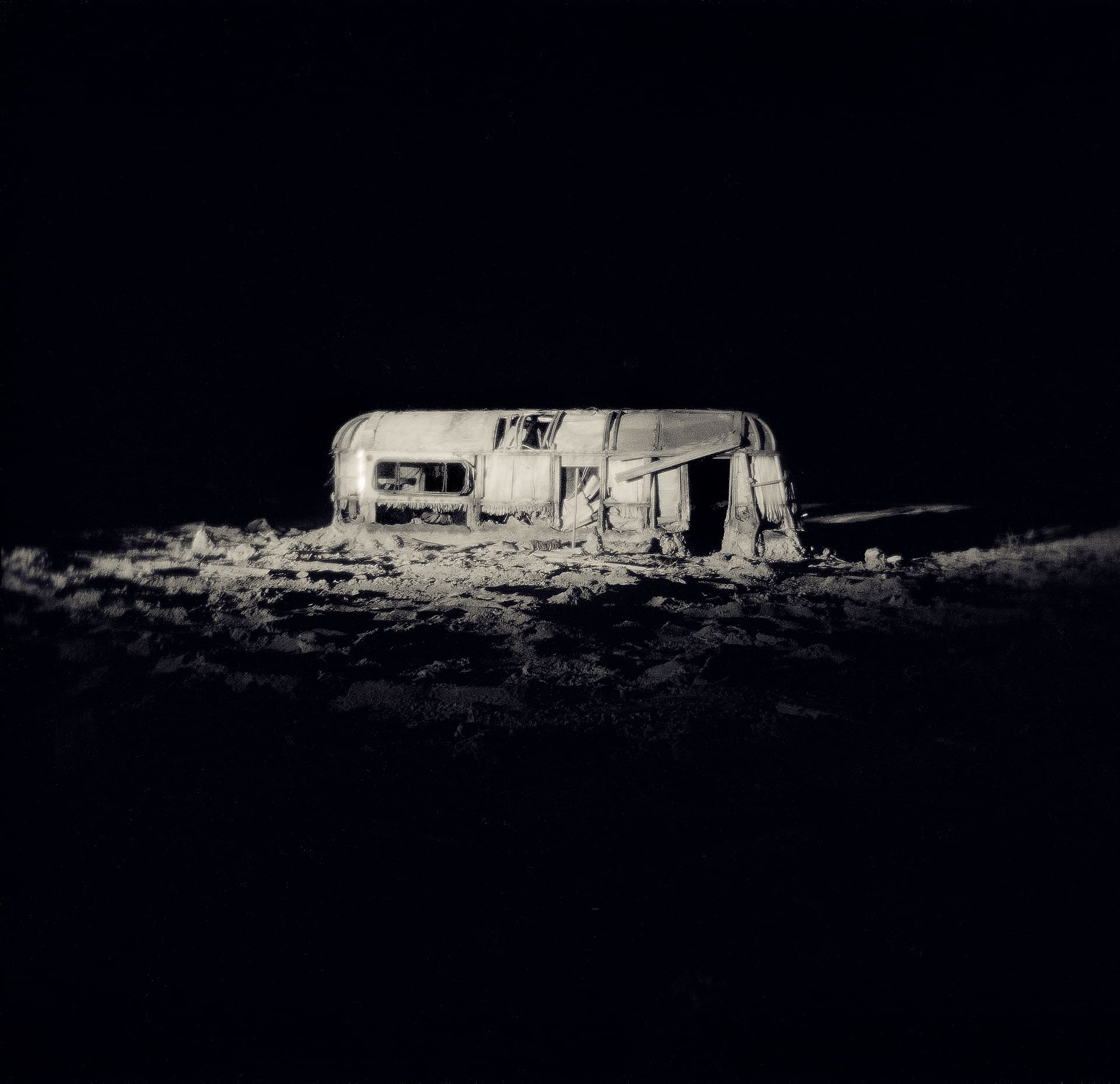 Wrecked caravan lit alone at night against total darkness, its shell and the ground around it glowing