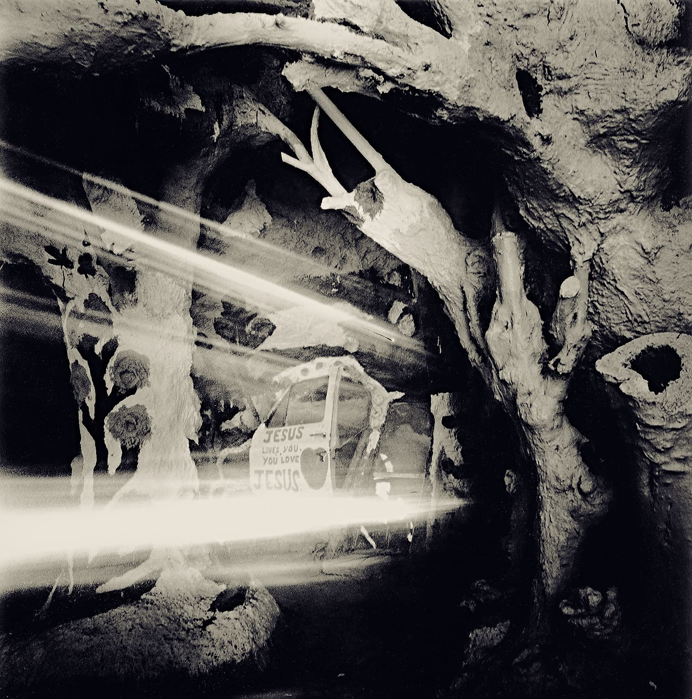 Painted adobe cave interior with sculpted tree trunks, a truck door reading Jesus loves you, and light streaks