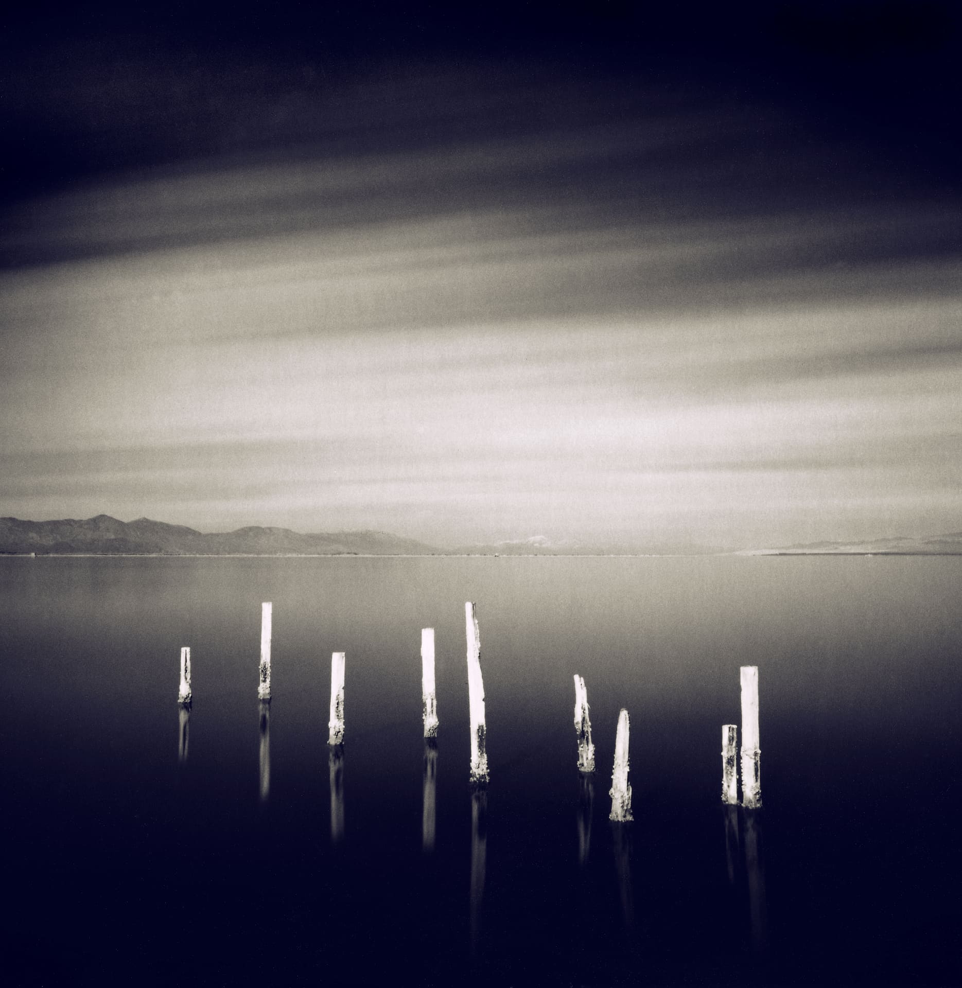 Nine pale broken posts standing in glassy dark water, low mountain ridge and streaked cloud beyond