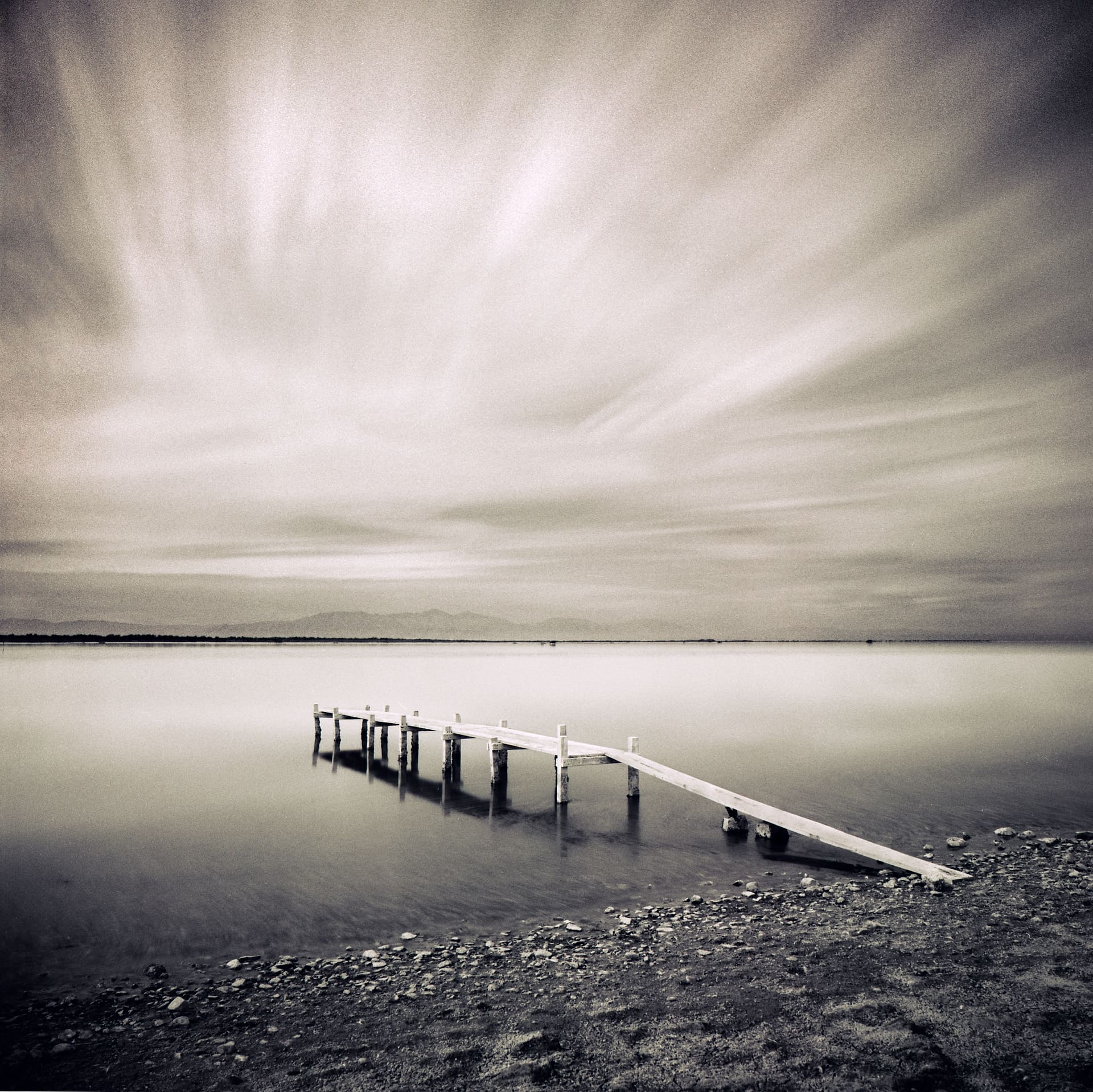 Collapsing wooden jetty leaning into flat water from a stony shore, long exposure smoothing the sea and streaking cloud