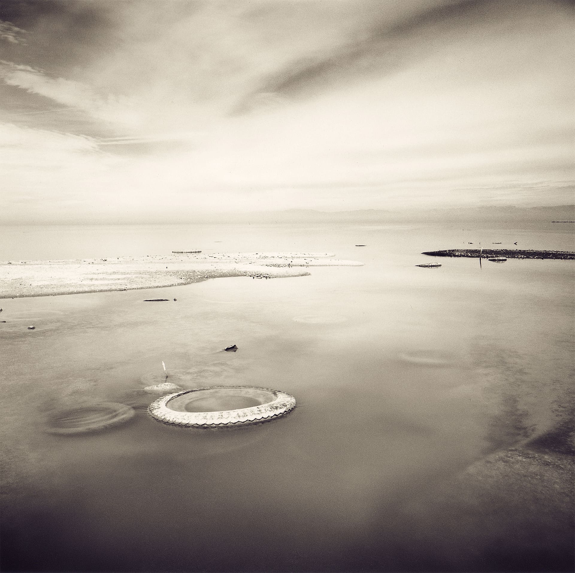Single tyre half-submerged in glassy shallow water, salt-crusted spit and distant mountains beyond, streaked cloud