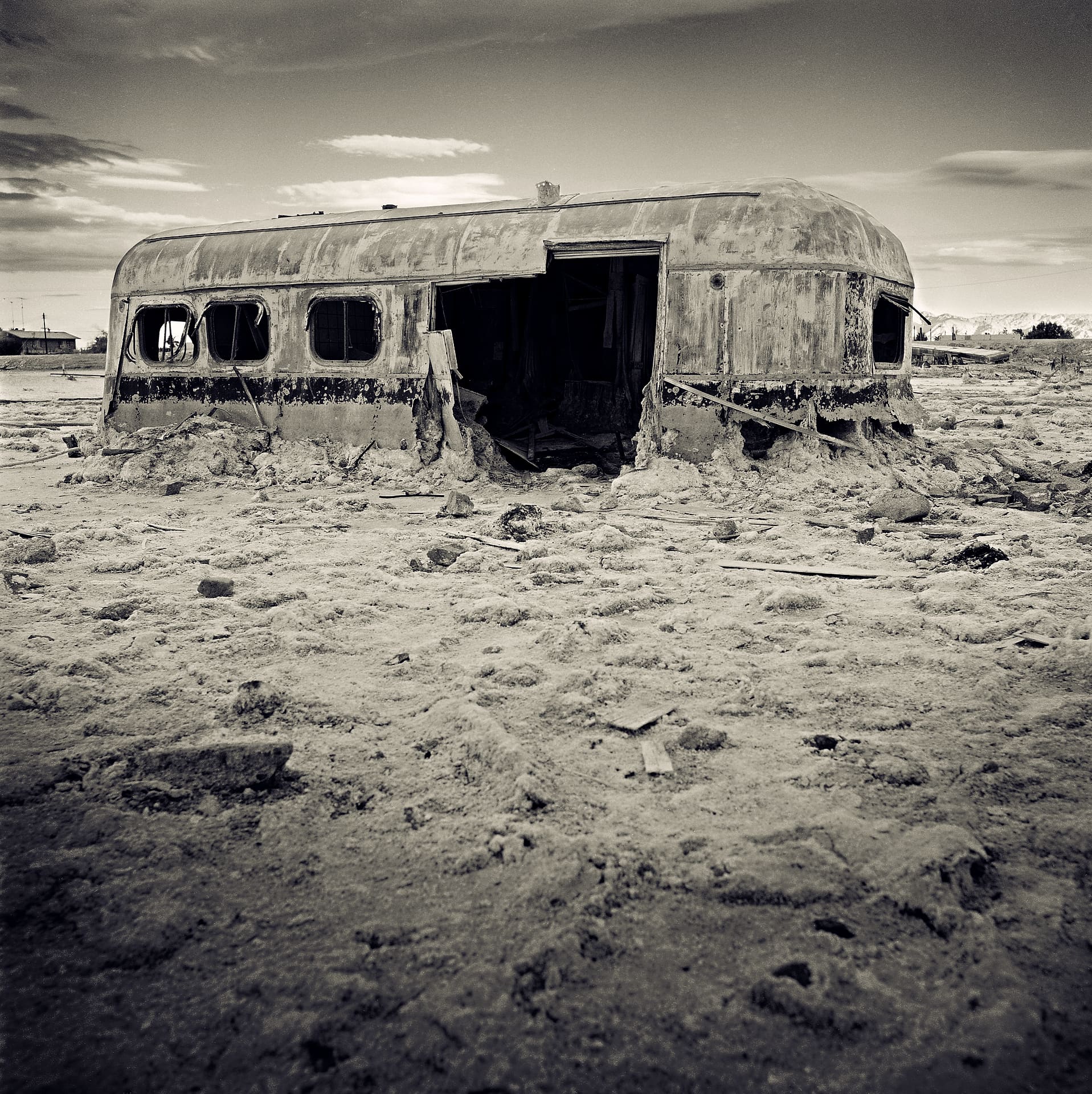 Rusted caravan with open doorway and broken windows sunk into a crusted salt flat scattered with debris