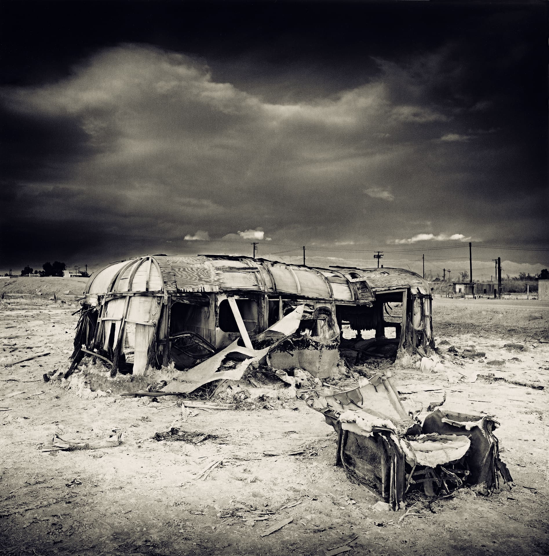 Burnt and flattened caravan shell on cracked salt flats, a torn car seat lying in the foreground