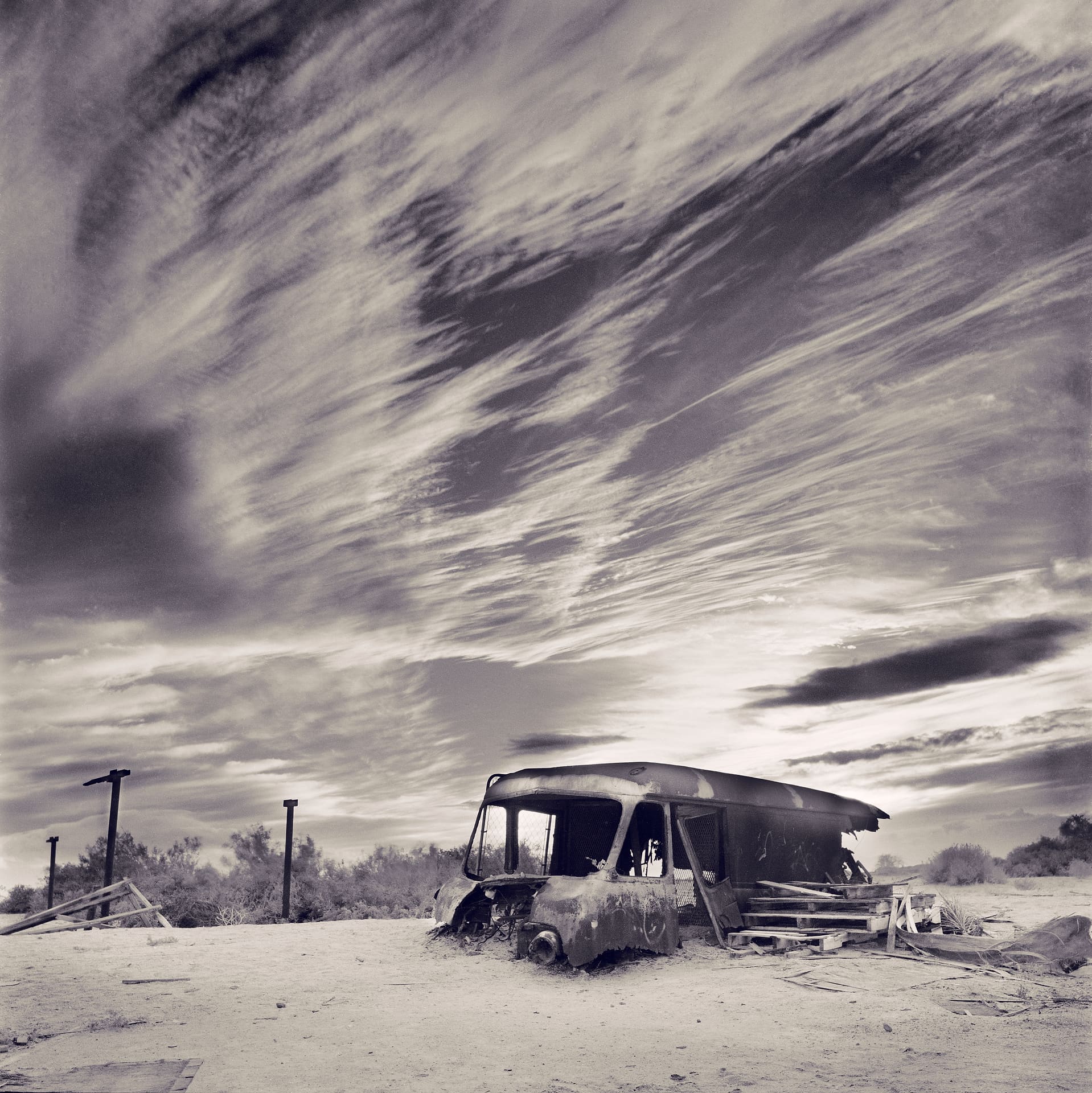 Burnt-out gutted van on sandy ground with scattered timber pallets, desert scrub and streaked cirrus cloud overhead