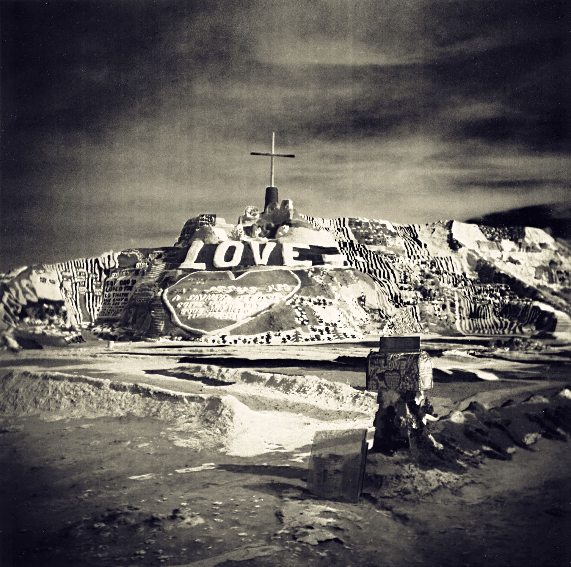 Painted adobe hillside with LOVE lettering and a cross on its summit, desert ground in the foreground