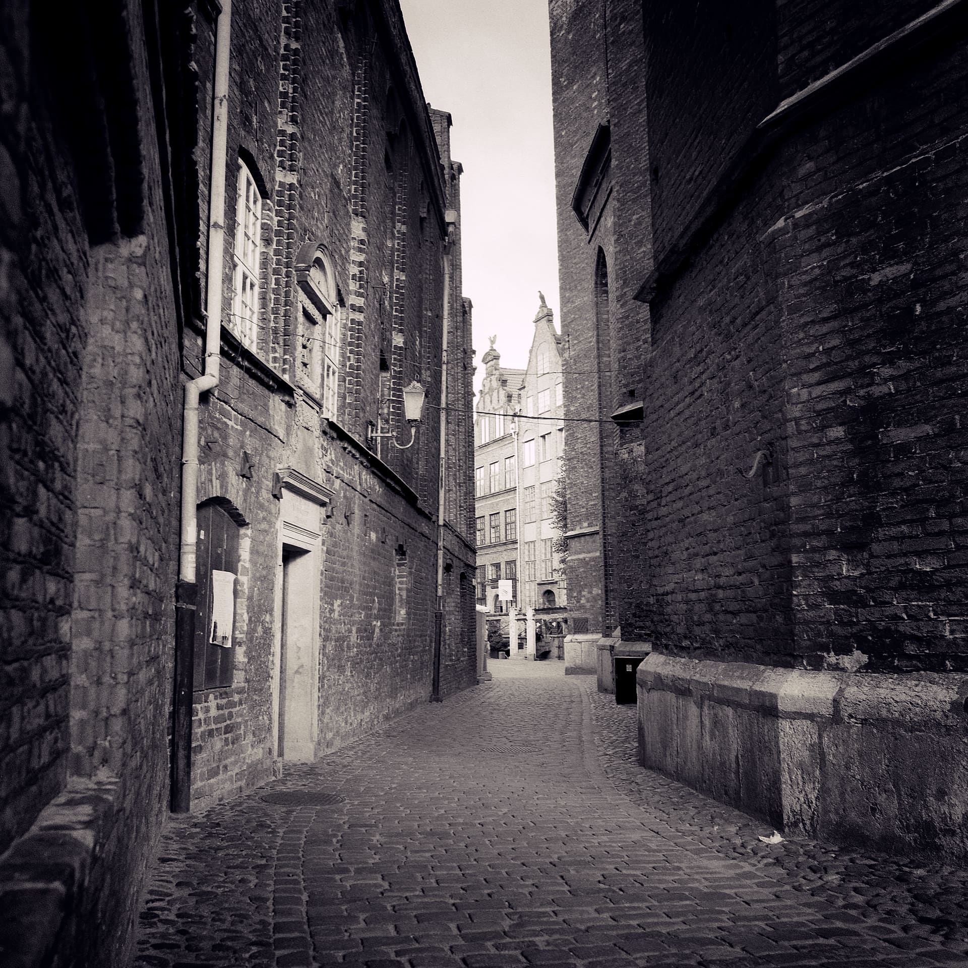 Narrow cobbled alley between tall brick walls opening onto a gabled townhouse and church at the far end
