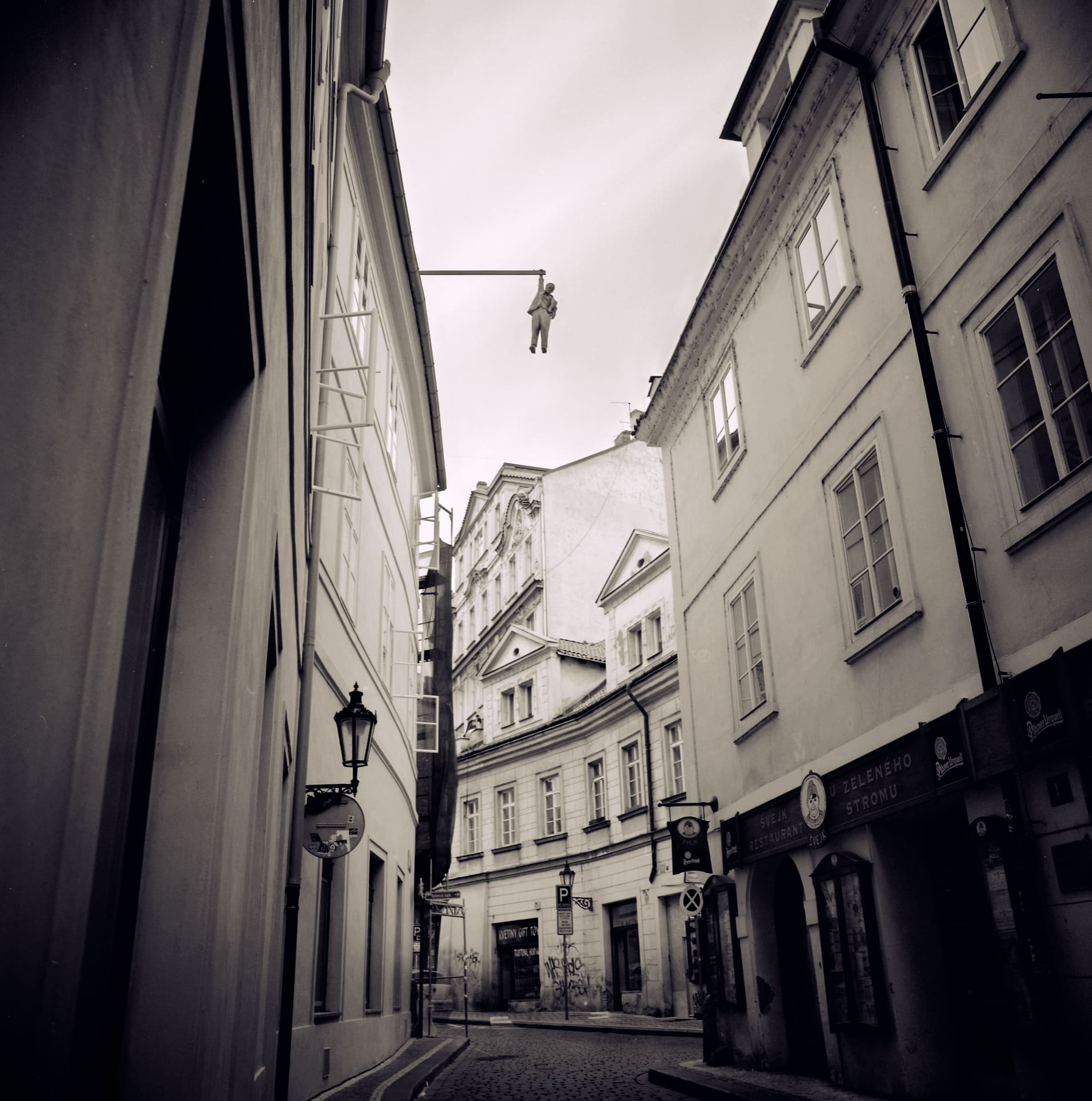 Empty cobbled street between tall plastered houses, a sculpture of a hanging man suspended from a pole overhead