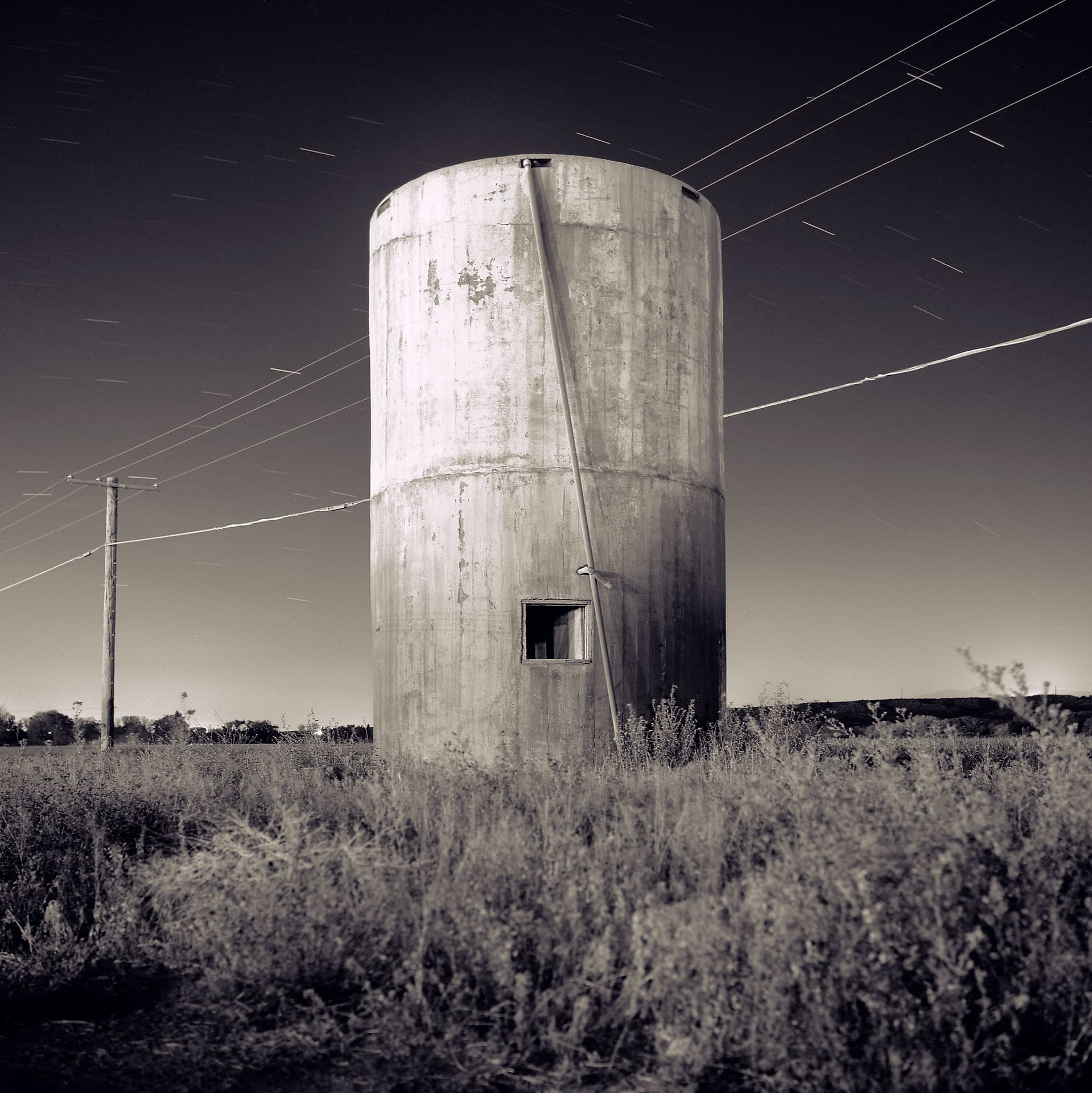 Concrete silo lit at night in weedy field, power lines overhead and short star trails streaking the sky