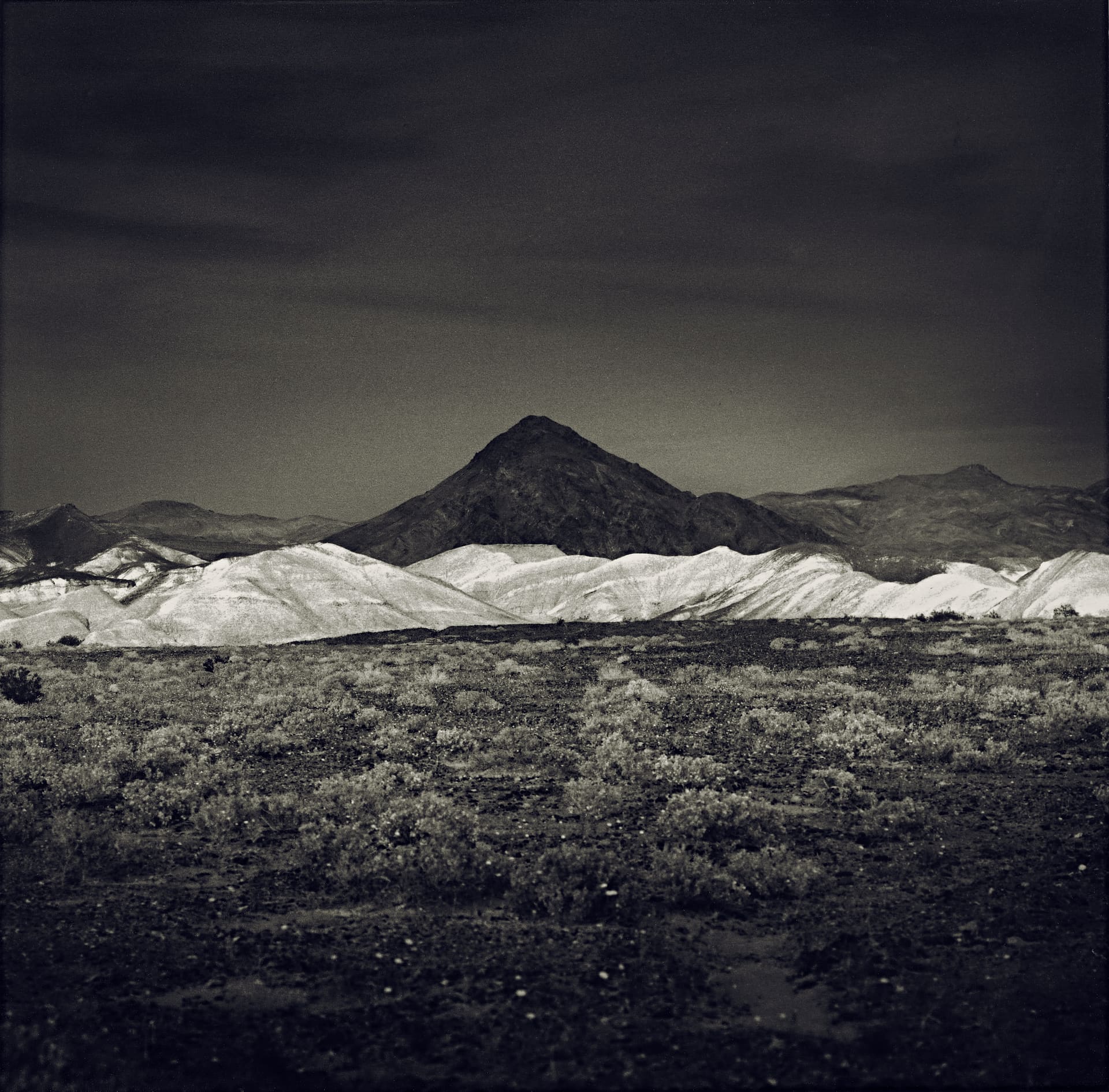 Dark conical peak above pale eroded badland ridges, desert scrub in the foreground under a heavy sky