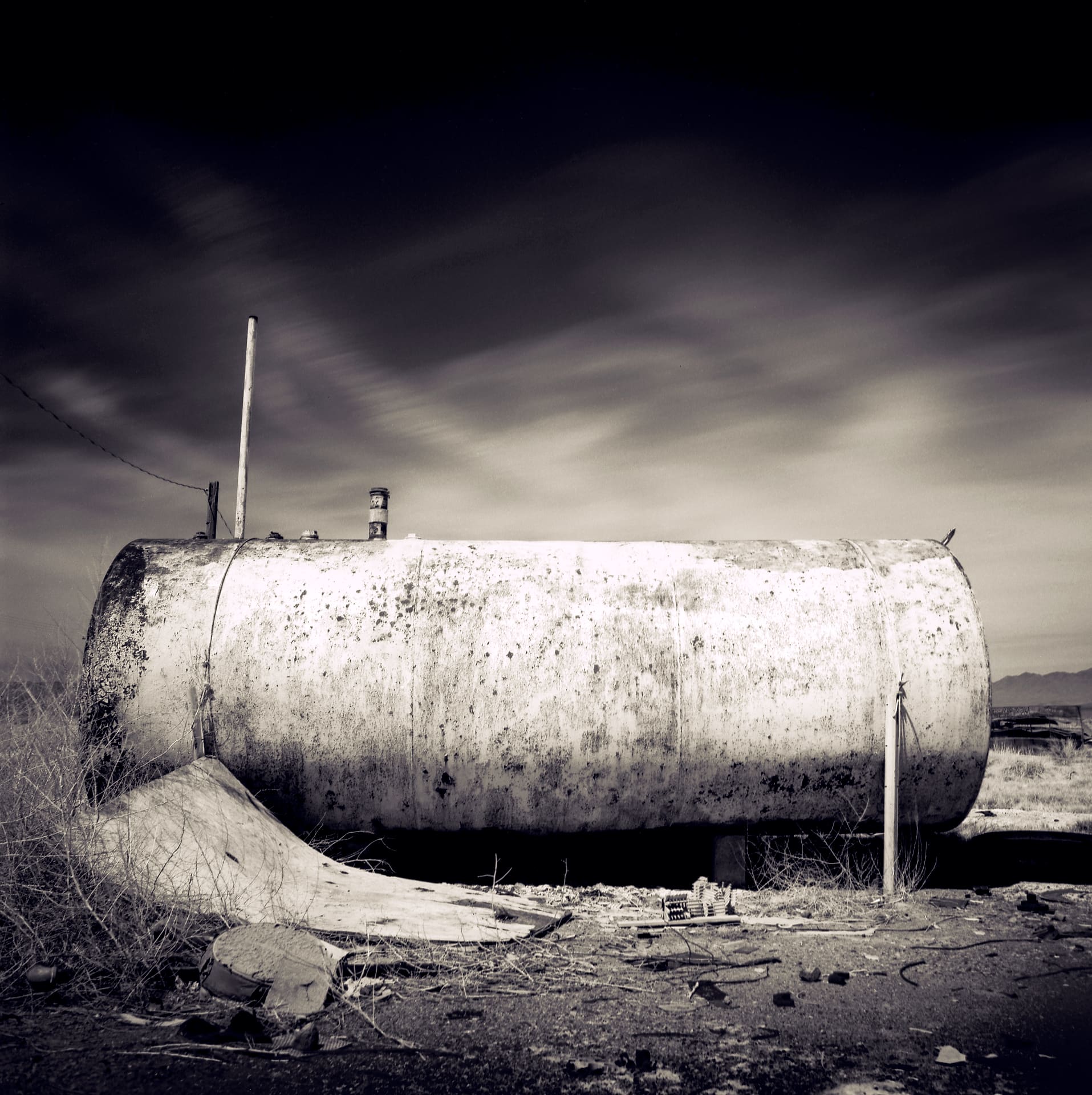 Weathered cylindrical fuel tank on cracked ground with scattered debris and dry brush, long exposure streaking cloud