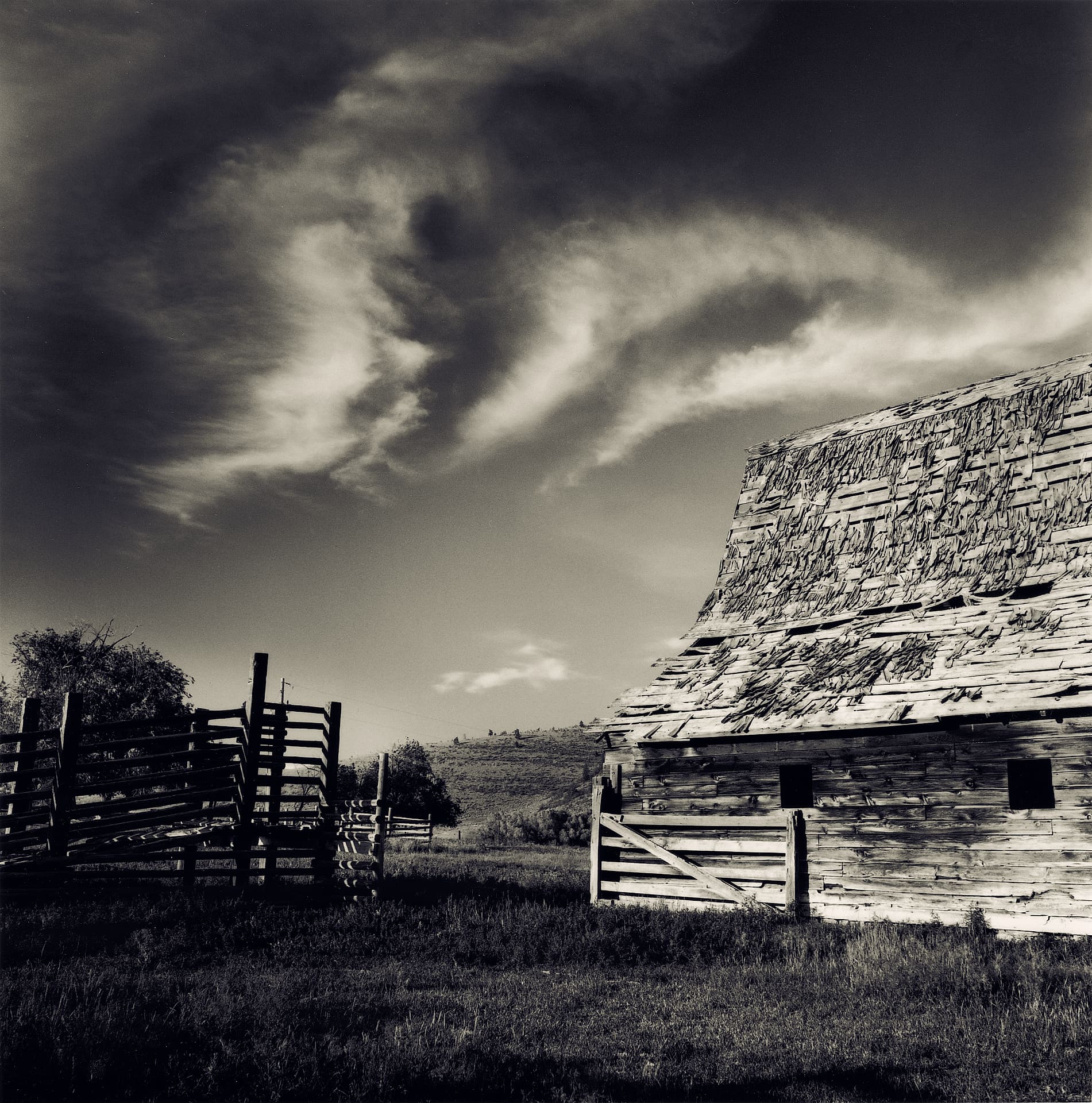 Weathered clapboard barn with sagging shingle roof beside wooden stock corrals under wispy cirrus cloud