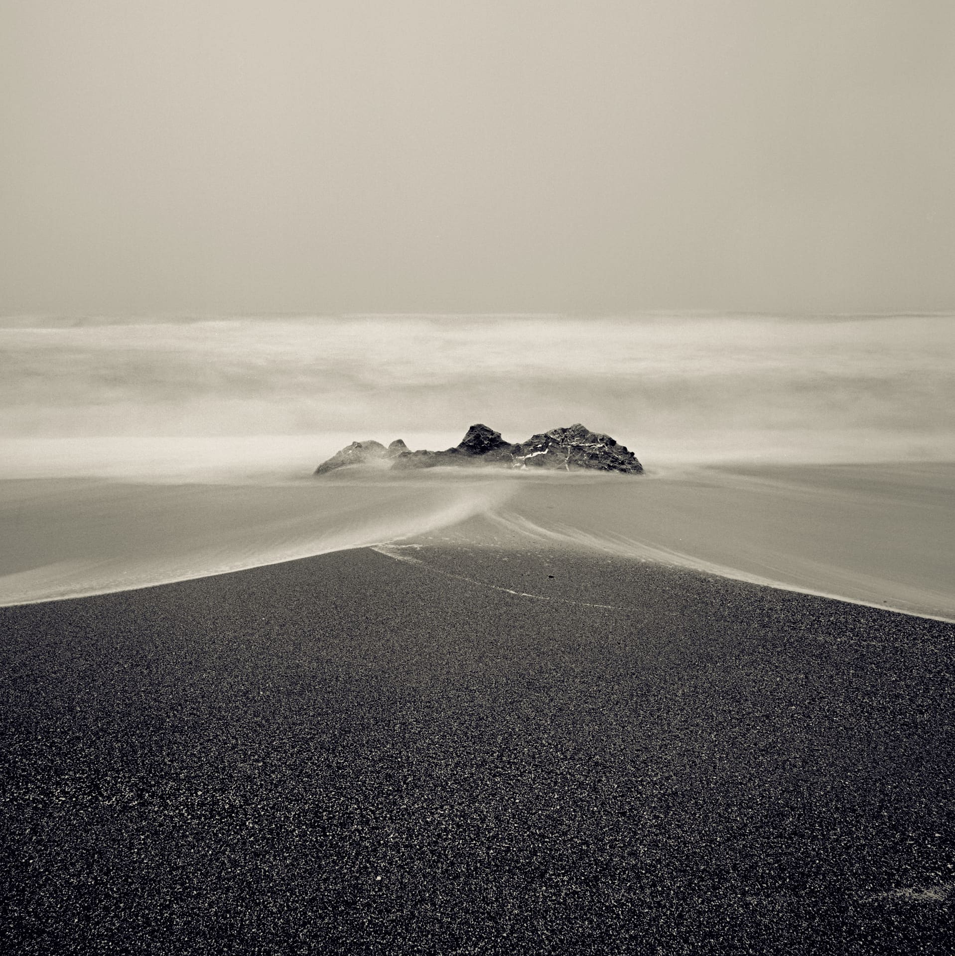 Small rock rising from surf smoothed to mist by long exposure, dark sand berm sweeping across foreground