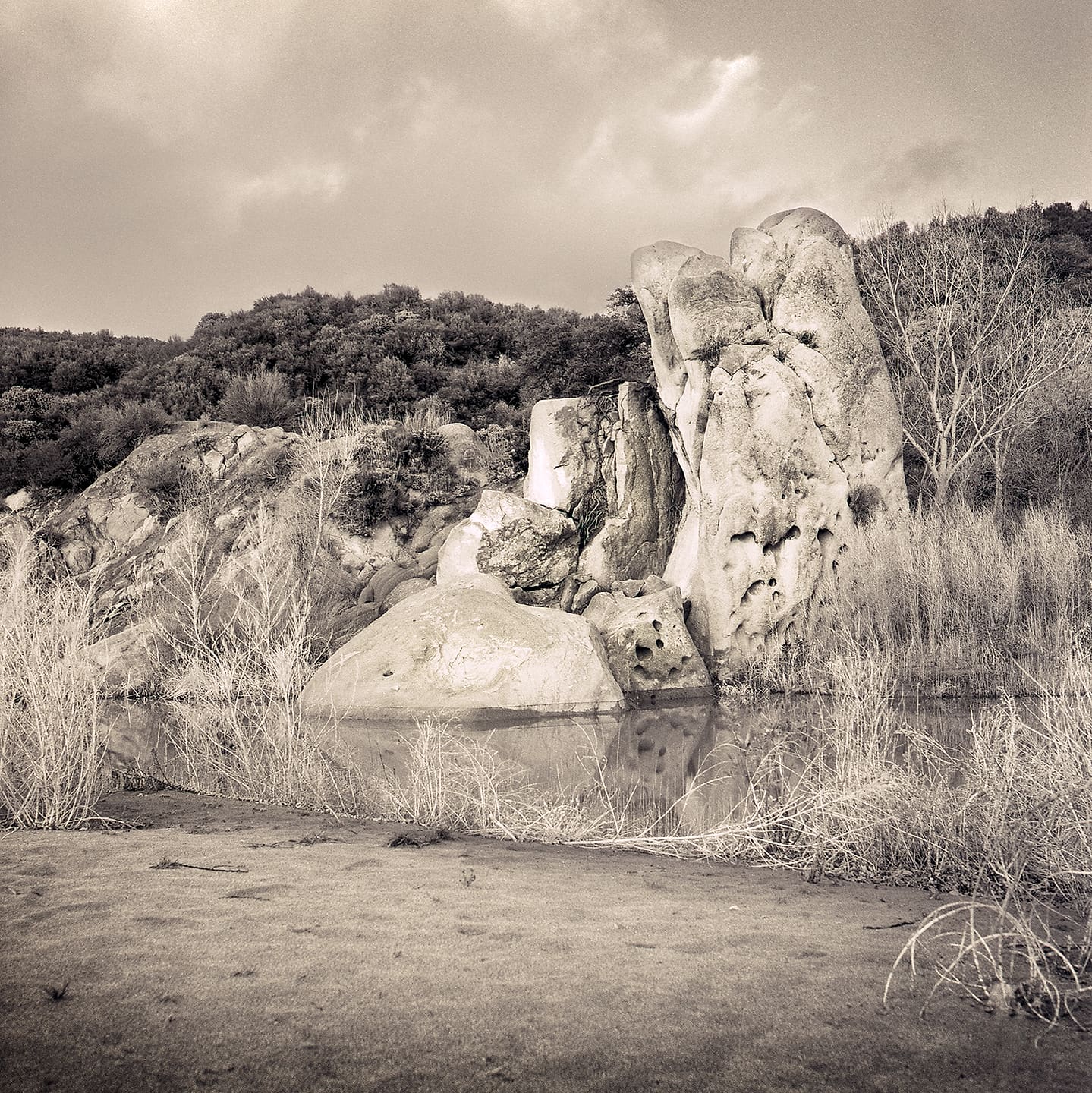 Weathered white boulders rising from a still shallow pool, dry brush and brooding cloud above