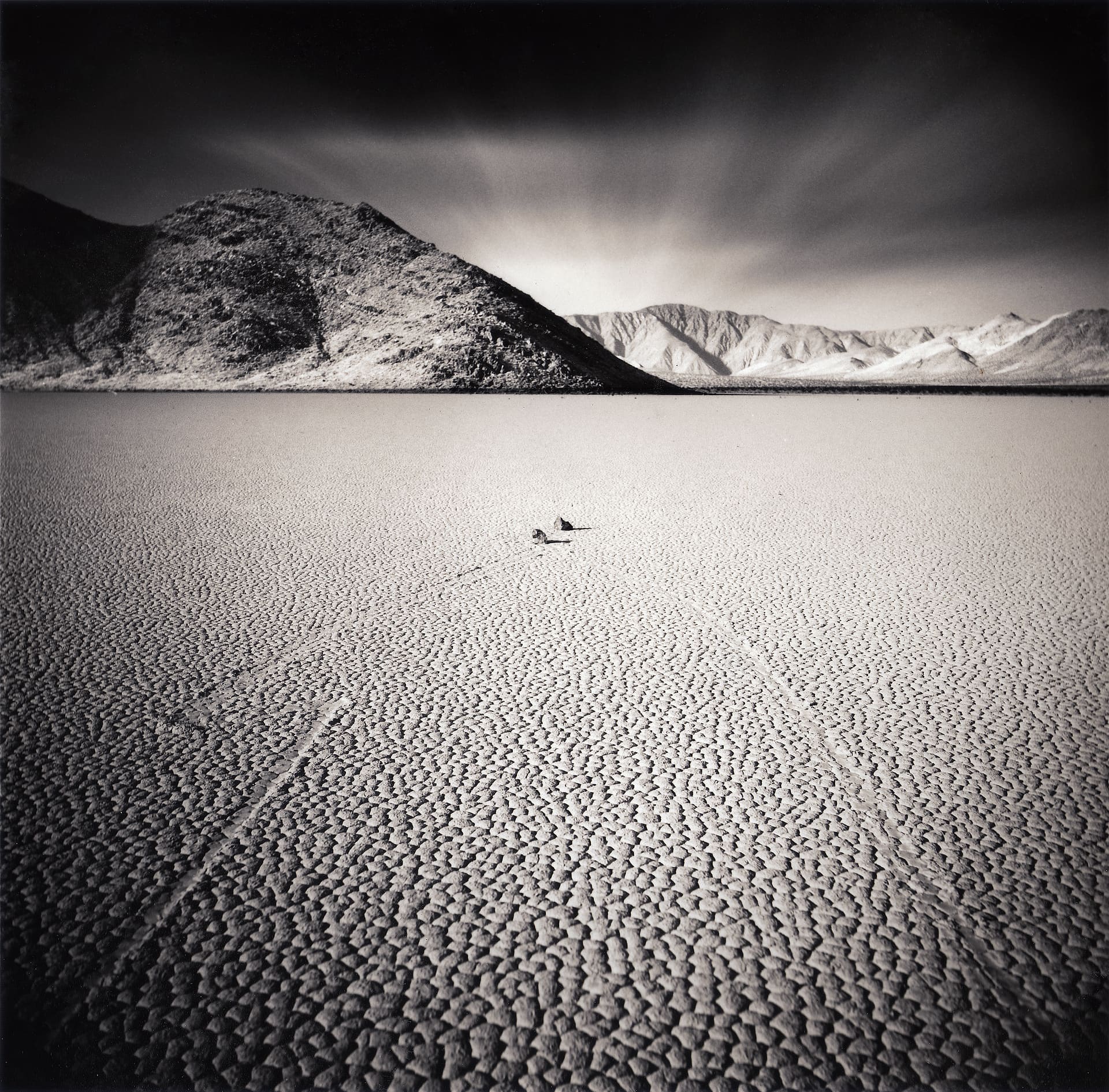 Cracked dry lakebed with a sailing stone and its long track, dark mountains and streaked cloud above
