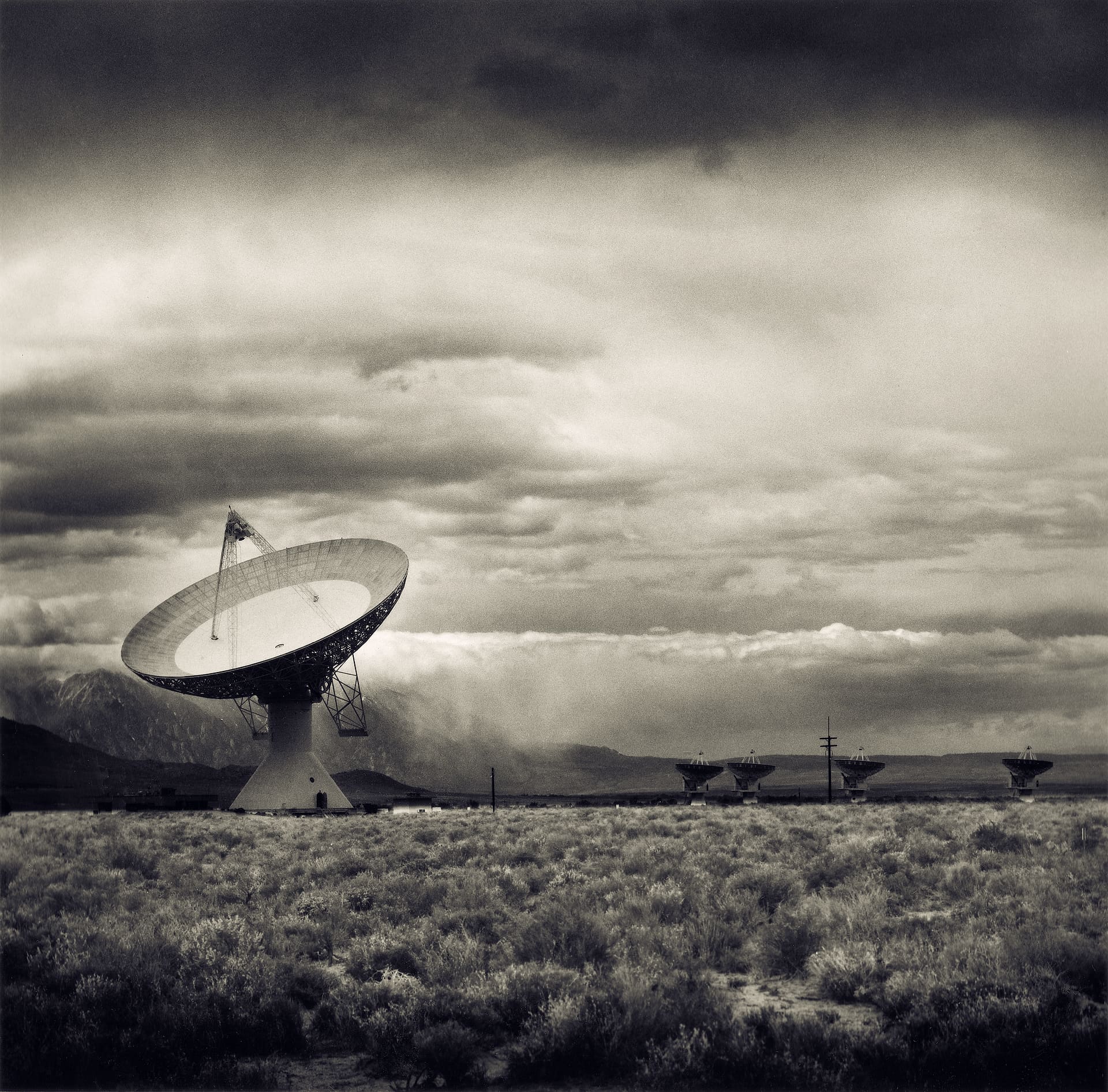 Large parabolic radio dish on sagebrush plain with smaller dishes in a line beneath heavy storm cloud