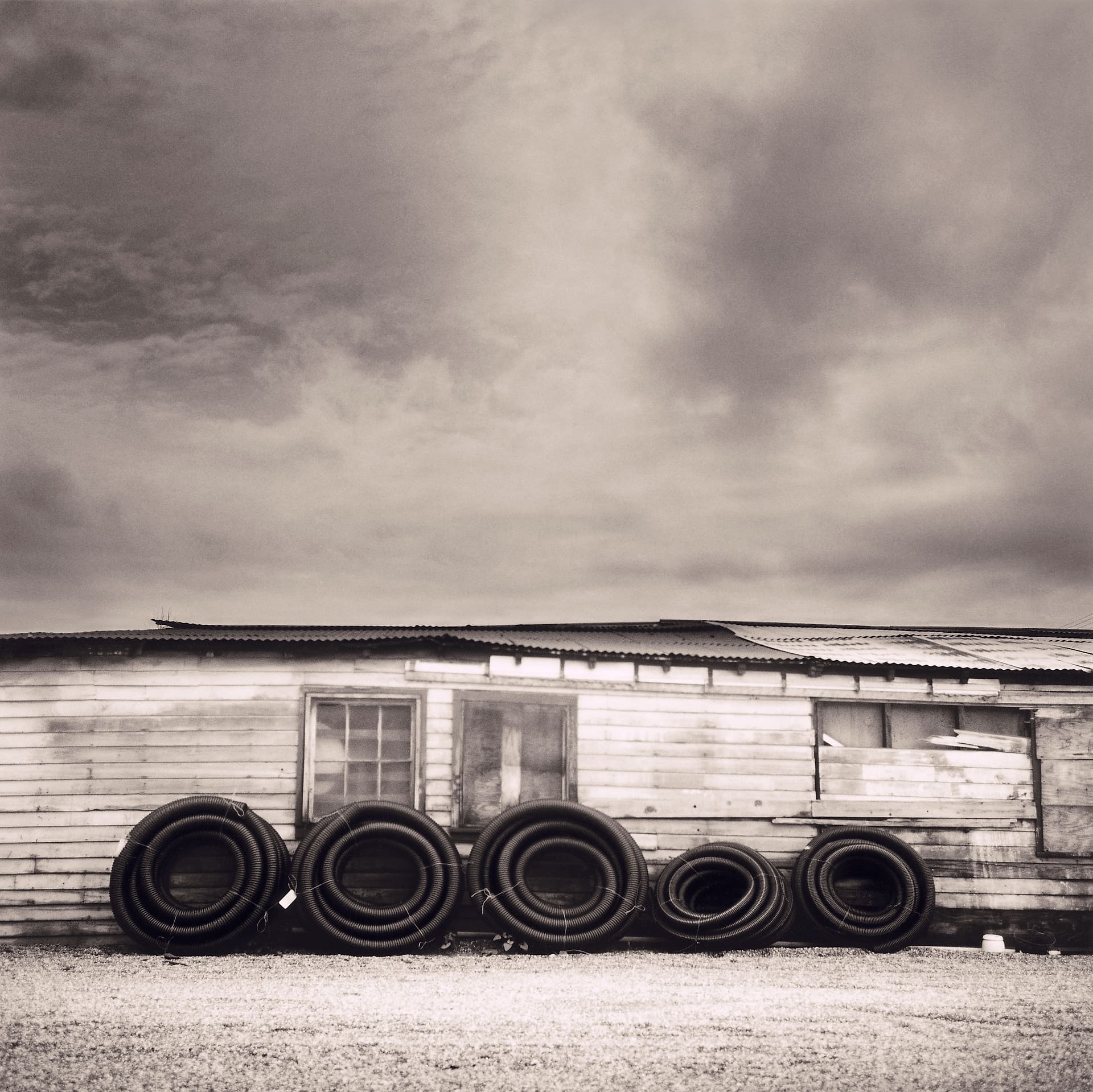 Five coils of black corrugated pipe stacked against a peeling clapboard shed on gravel under grey cloud