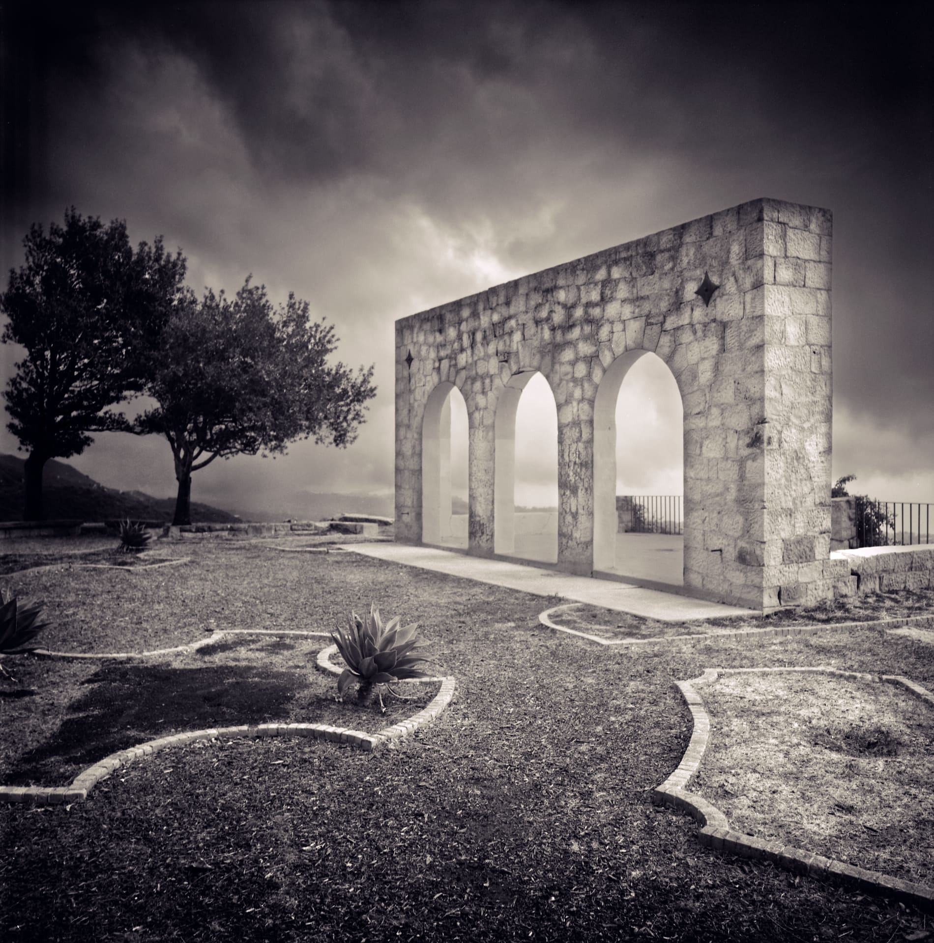 Freestanding stone wall pierced by three arches on a clipped hillside garden, storm cloud massing behind